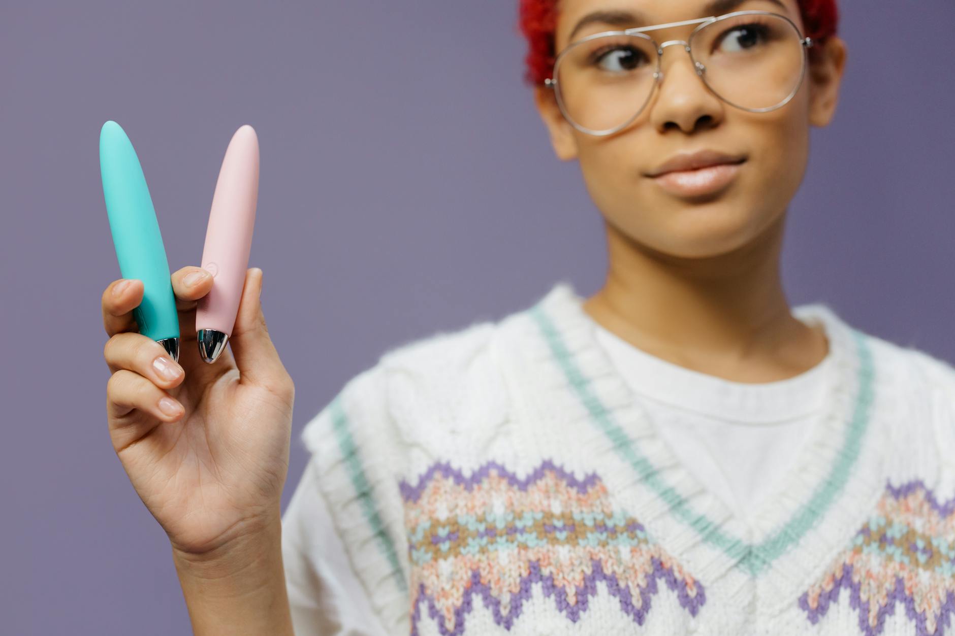 Mulher jovem segurando dois vibradores coloridos contra fundo roxo